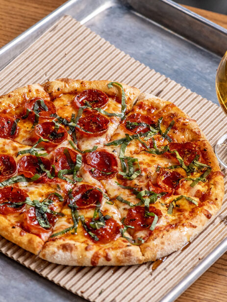 Hot Pepperoni Pizza from Ramundos at Element Eatery food hall, topped with fresh basil, served with Ramundos Bites, marinara sauce, beer, and peach vodka seltzer on a wooden table