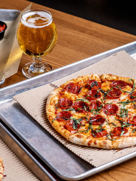 Hot Pepperoni Pizza from Ramundos at Element Eatery food hall, topped with fresh basil, served with Ramundos Bites, marinara sauce, beer, and peach vodka seltzer on a wooden table