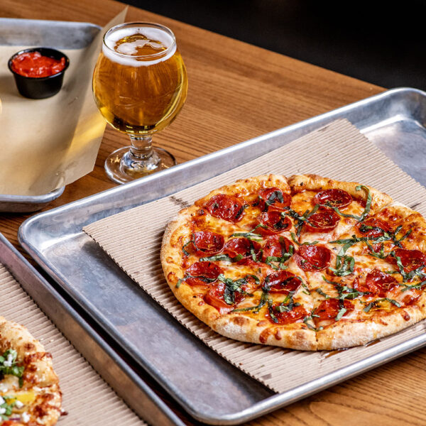 Hot Pepperoni Pizza from Ramundos at Element Eatery food hall, topped with fresh basil, served with Ramundos Bites, marinara sauce, beer, and peach vodka seltzer on a wooden table