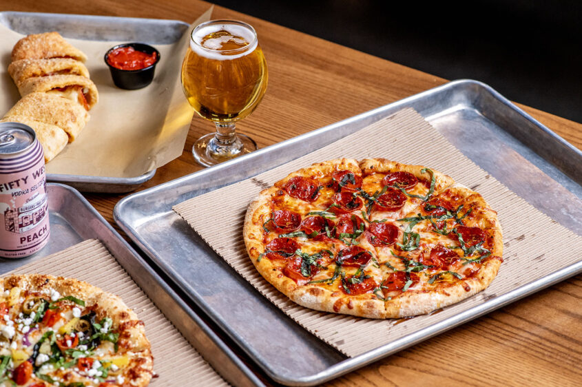 Hot Pepperoni Pizza from Ramundos at Element Eatery food hall, topped with fresh basil, served with Ramundos Bites, marinara sauce, beer, and peach vodka seltzer on a wooden table