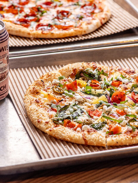 Calypso Pizza from Ramundos at Element Eatery food hall, topped with fresh vegetables and cheese, served with a peach vodka seltzer on a metal tray