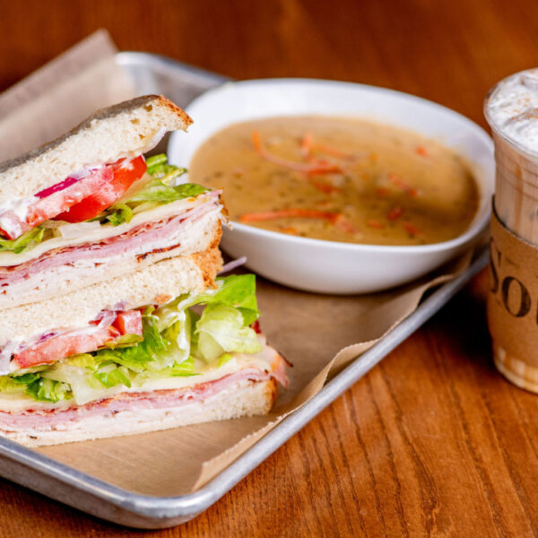 Stacked deli sandwich with lettuce, tomato, and sliced meats, served with a bowl of soup and iced drink from Solare Café at Element Eatery food hall