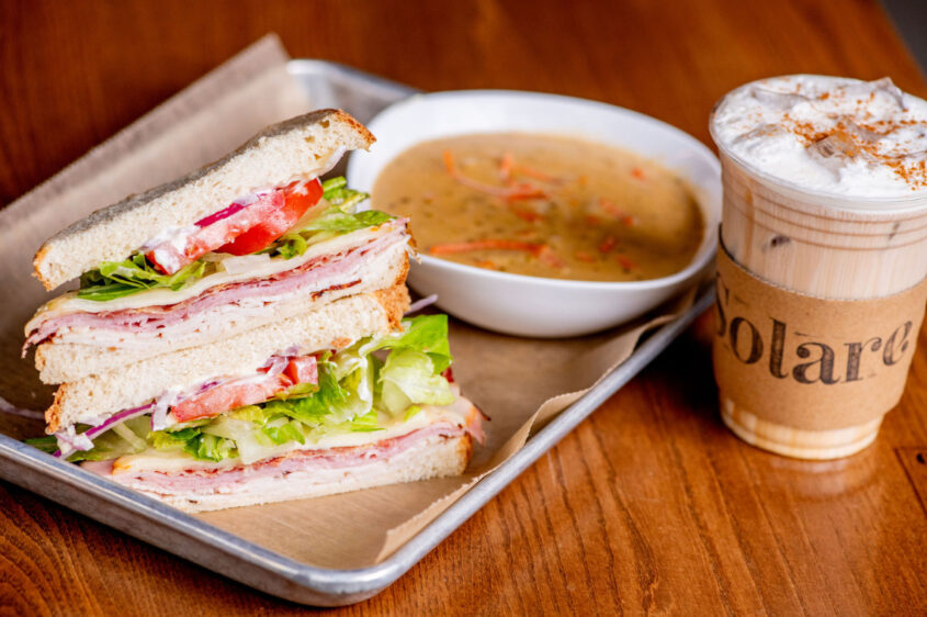 Stacked deli sandwich with lettuce, tomato, and sliced meats, served with a bowl of soup and iced drink from Solare Café at Element Eatery food hall