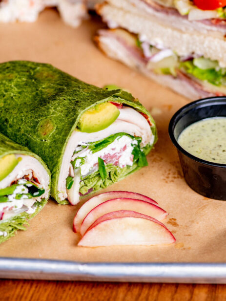 Turkey avocado spinach wrap with sliced apples and dipping sauce from Solare Café at Element Eatery food hall, served on a metal tray with brown parchment paper