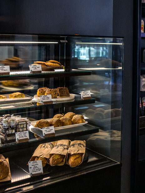 Bakery case at Solare Café in Element Eatery food hall, displaying fresh breads, pastries, and baked goods alongside a refrigerated wall of canned and bottled drinks