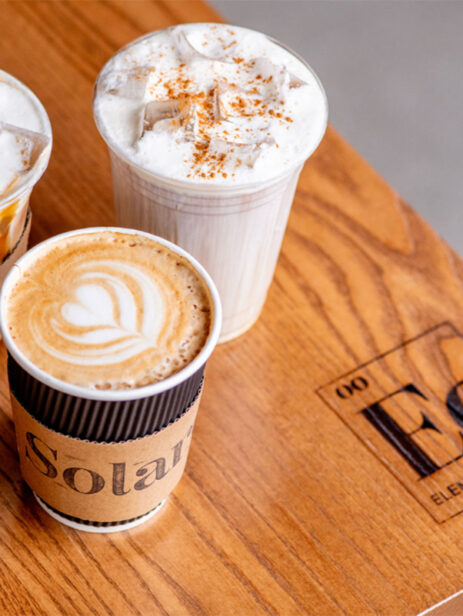 Coffee drinks from Solare Café at Element Eatery food hall, featuring a hot latte with latte art and two iced drinks topped with whipped cream and caramel drizzle
