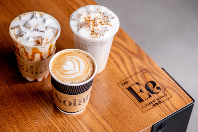 Coffee drinks from Solare Café at Element Eatery food hall, featuring a hot latte with latte art and two iced drinks topped with whipped cream and caramel drizzle